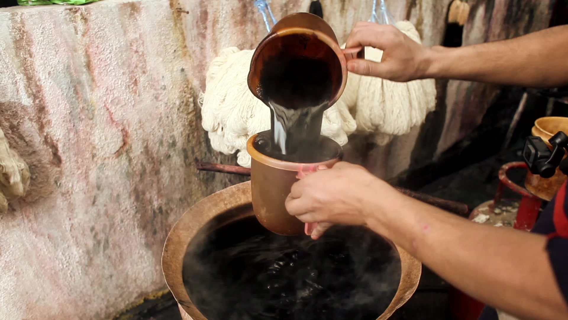 Dye poured from one copper pot into another beside a dyeing vat, with pale skeins of yarn hanging behind