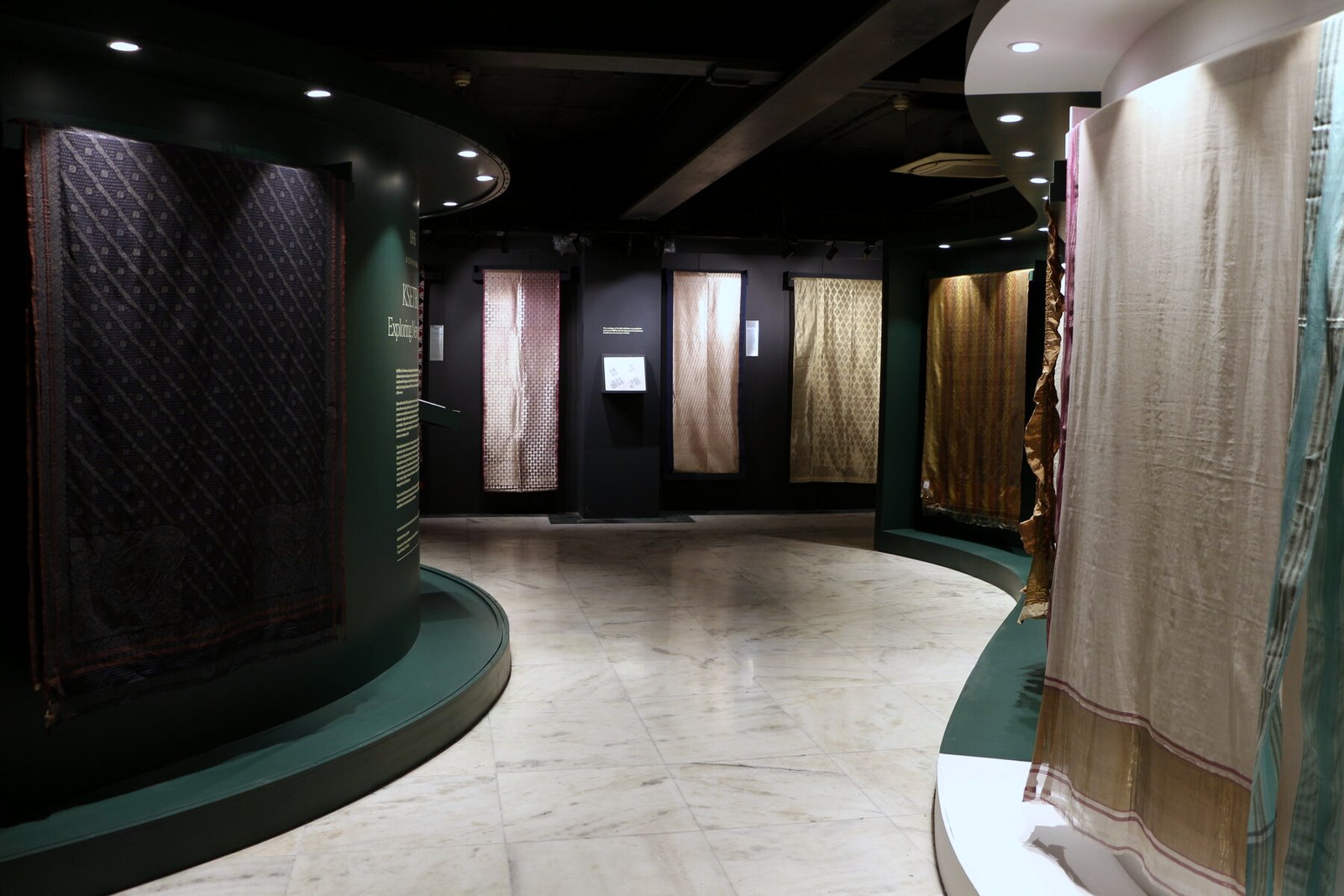 Gallery interior with textile displays on curved green walls and illuminated panels.