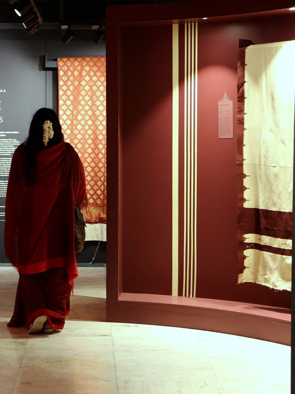 Woman in red sari viewing display case with orange patterned textile in gallery.
