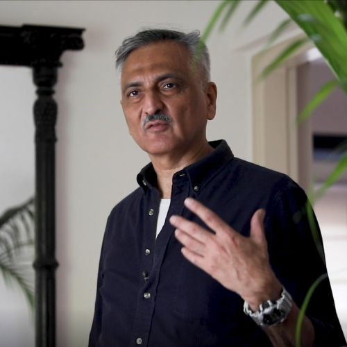 Rakesh Thakore in a dark shirt, speaking, beside a carved wooden column