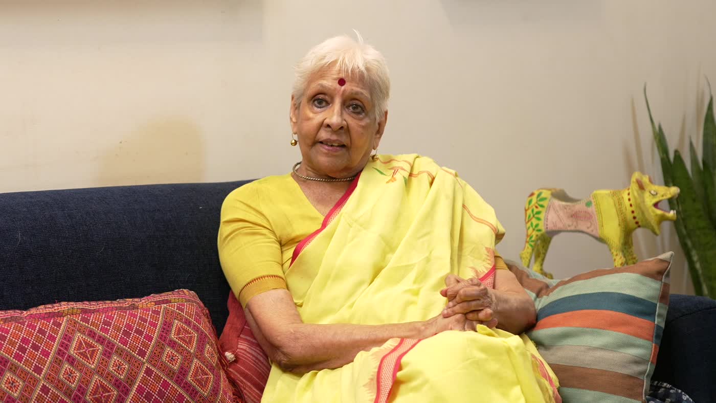 Jaya Jaitly seated on a sofa in a yellow handwoven sari, with handloom cushions and a painted wooden figure beside her
